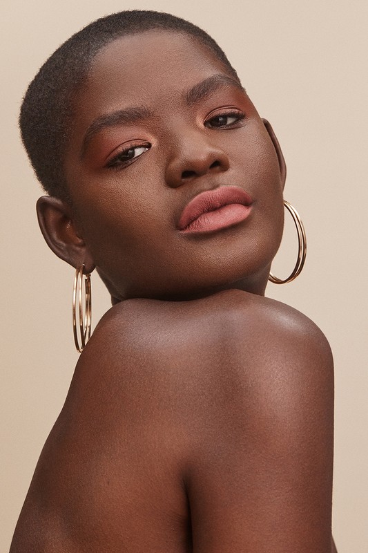 Close-up shot of a beautiful dark-skinned woman with short hair, wearing gold hoop earrings and makeup, looking confidently at the camera.