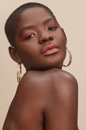 Close-up beauty portrait of a Black woman with short hair wearing large gold hoop earrings against a neutral beige background