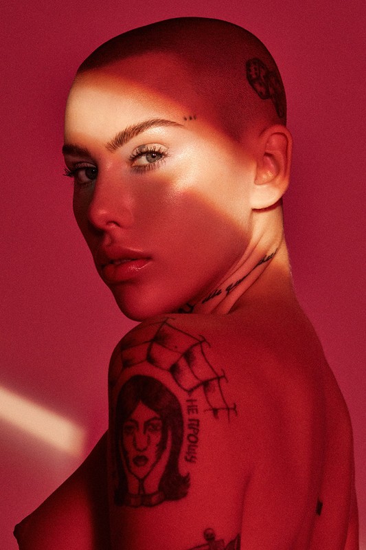 Close-up portrait of a woman with a shaved head against a red background, featuring tattoos on her shoulder and neck.