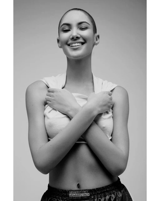Portrait of a smiling woman with a shaved head, wearing a tank top with arms crossed, in a black and white studio setting.