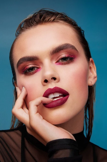 Close-up studio shot of a young adult woman with wet hair, bold maroon lipstick, and dramatic pink and maroon eye makeup against a blue background.