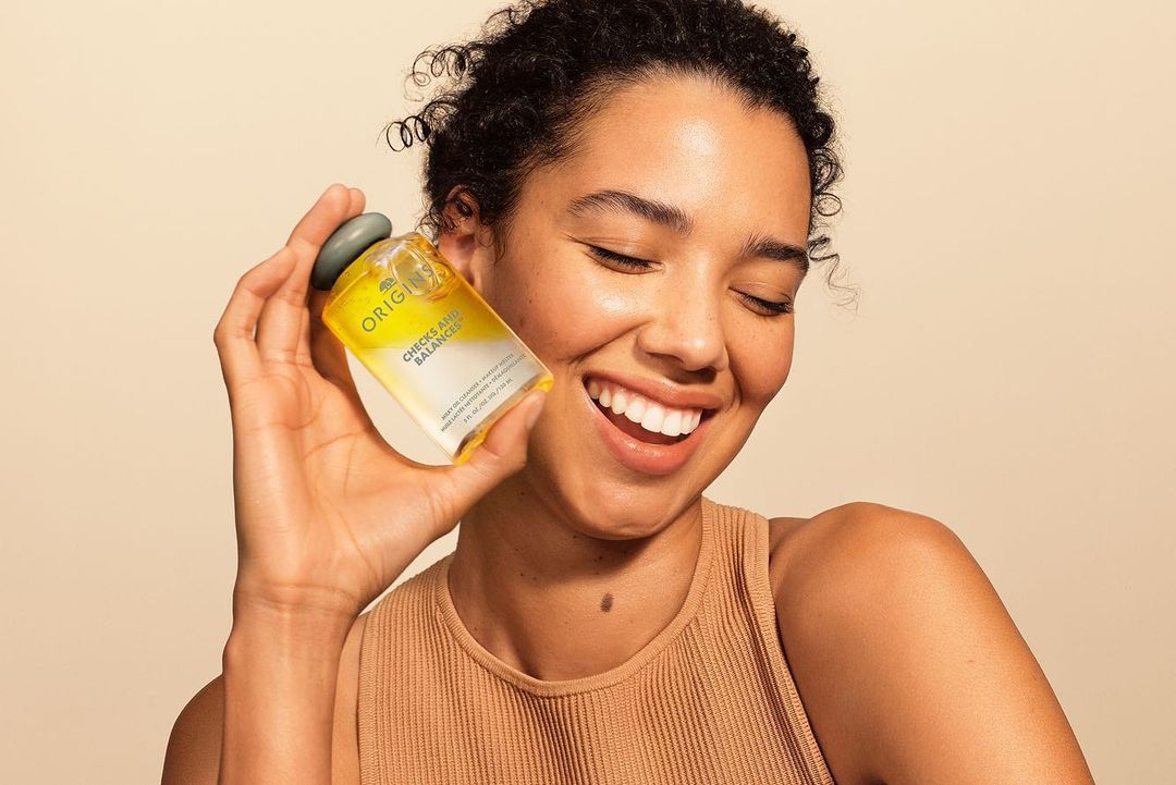 Smiling woman holding bottle of Origins Checks and Balances face cleanser. Beauty shot with soft, neutral background. Skincare product.
