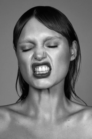 Close-up monochrome portrait of a woman with wet hair scrunching her face and baring teeth against a plain grey background