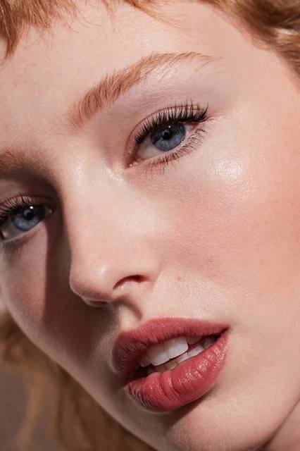 Close-up portrait of a fair-skinned young woman with red hair, blue eyes, and light pink lipstick, featuring soft lighting and a blurred background.