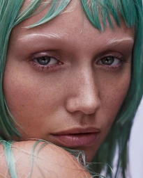 Close-up portrait of a woman with vibrant teal hair and bleached eyebrows looking directly at the camera.