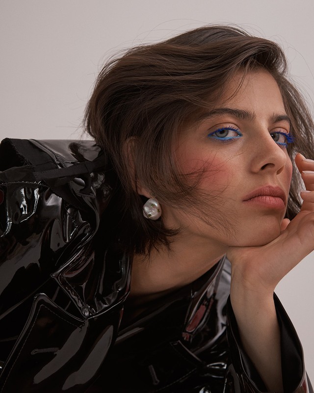 Close-up portrait of a woman with short dark hair, wearing a black patent leather jacket and statement silver earrings against a neutral backdrop.