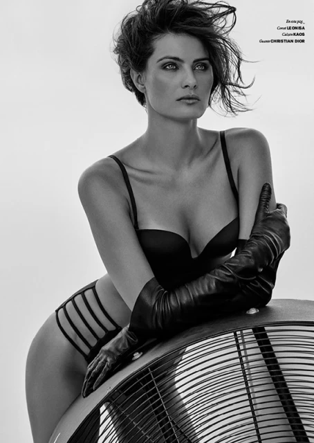 Monochrome portrait of an adult female model in black lingerie and gloves, leaning on a fan against a plain white background. Studio lighting.