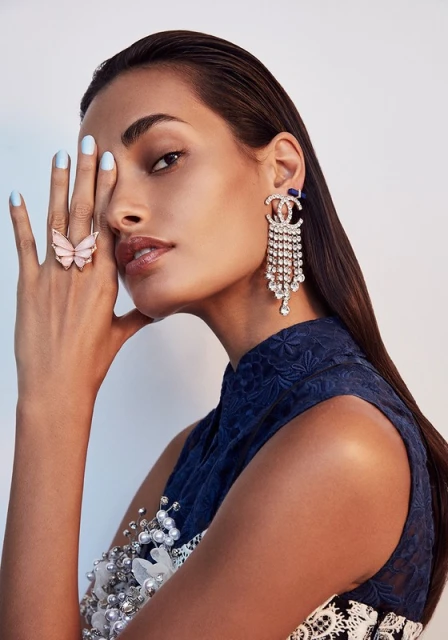 Portrait of a woman with slicked-back dark hair wearing a blue patterned dress, dangling crystal Chanel earrings, and a pink butterfly ring.