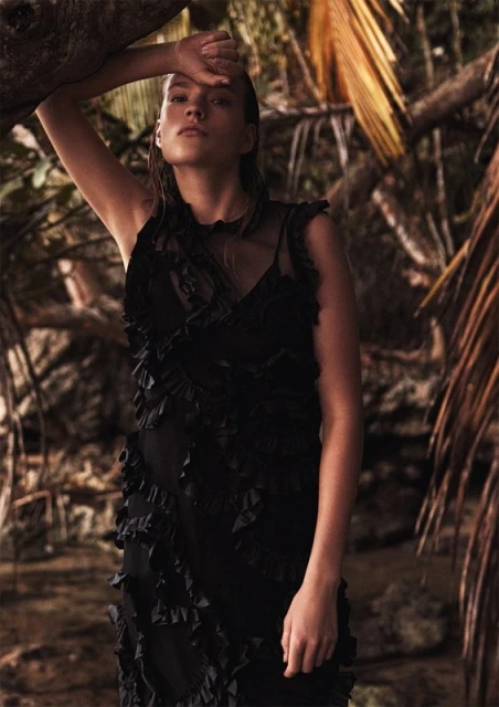 Woman in a black dress poses in a lush tropical forest setting, partially obscured by tree branches, with soft, diffused lighting.