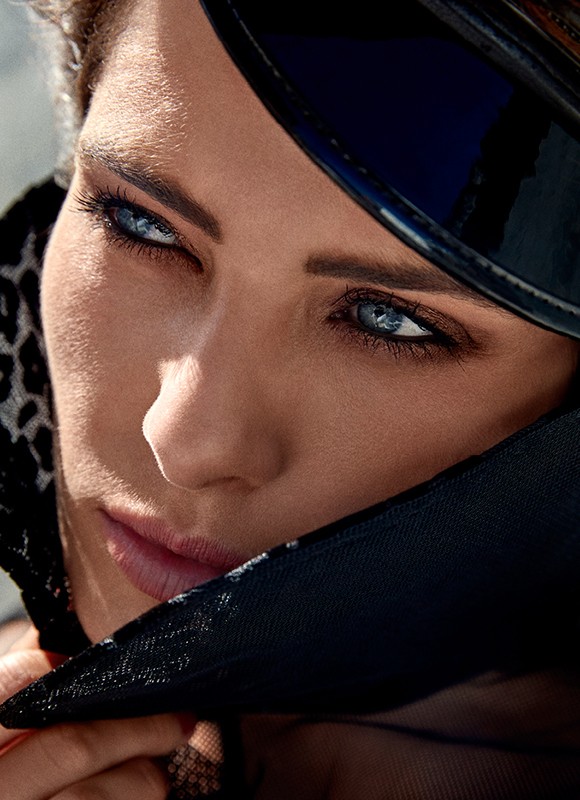 Close-up portrait of a woman with striking blue eyes, wearing a dark hat and sheer fabric, capturing a sense of mystery and allure.