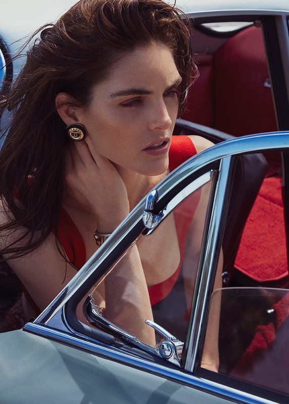 A woman with dark hair poses in a light blue vintage car. She wears a red top and gold earrings, with a blue sky in the background.