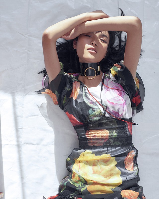 Eye-level shot of a young Asian woman in a floral dress lying on a white surface with arms crossed above her head, indoors.