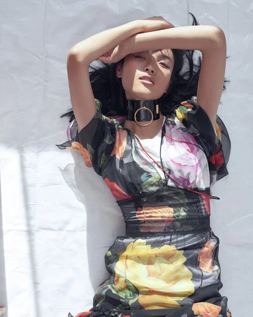 Eye-level shot of a young Asian woman in a floral dress lying on a white surface with arms crossed above her head, indoors.