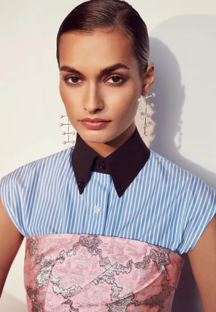 Close-up portrait of a woman wearing a blue striped collared shirt, pink patterned bustier, and pearl drop earrings.
