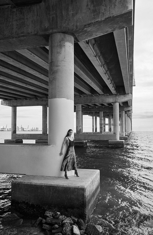 Monochrome shot of a woman in a dress posing beneath an elevated pier, with textured concrete pillars and ocean water.