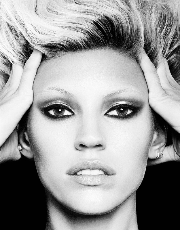Black and white close-up portrait of an adult female with bleached hair and dark eyeshadow, holding her hands on her head.