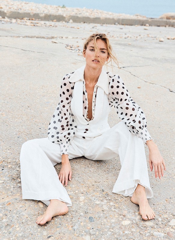 Elegant adult female in white polka-dot blouse and white jumpsuit sitting on concrete, looking at the camera with neutral expression.