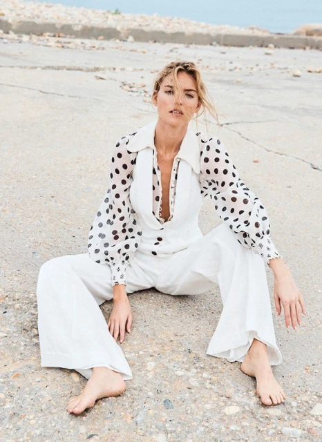 Elegant adult female in white polka-dot blouse and white jumpsuit sitting on concrete, looking at the camera with neutral expression.