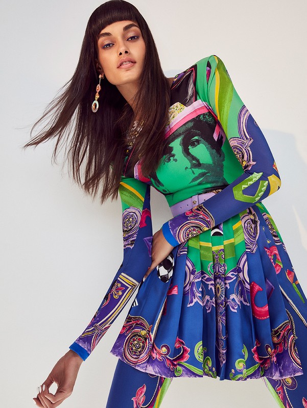 Brunette model wearing a multicolored patterned long-sleeve dress and mini-skirt in a studio setting, accessorized with large earrings.