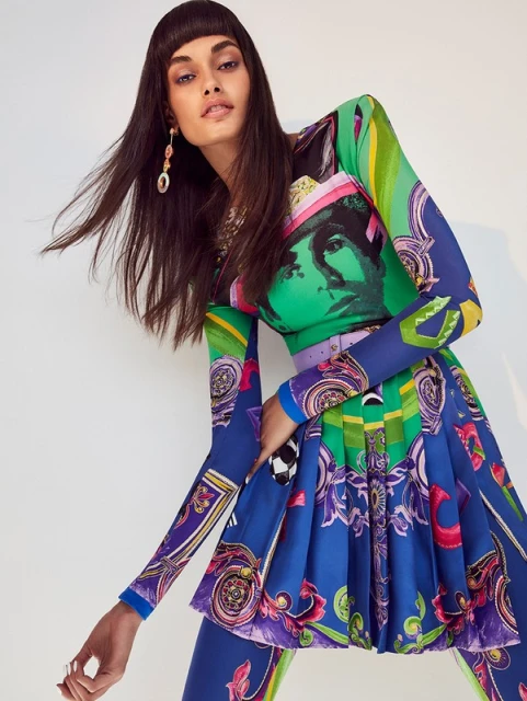 Brunette model wearing a multicolored patterned long-sleeve dress and mini-skirt in a studio setting, accessorized with large earrings. 