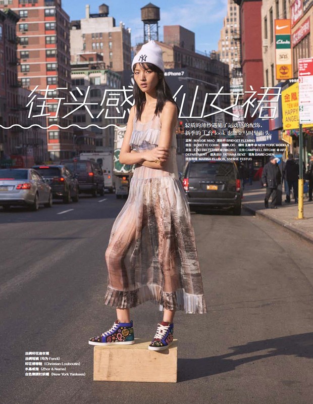 Fashion shot of an Asian woman wearing a long sheer dress, standing on a wooden box in the middle of a city street with traffic.