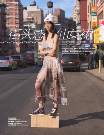 Fashion shot of an Asian woman wearing a long sheer dress, standing on a wooden box in the middle of a city street with traffic.