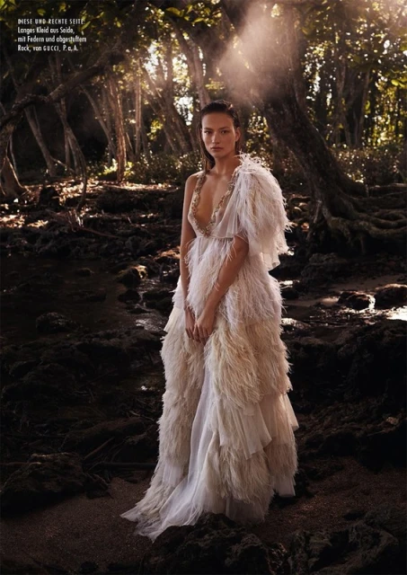 Woman in flowing, feathered gown poses in a sunlit forest. Light filters through trees, illuminating the scene for a fashion editorial.
