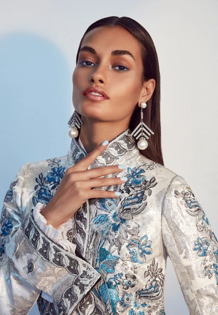 Portrait of a woman with fair skin and brown hair, wearing large pearl earrings and a floral top, set against a light blue background.