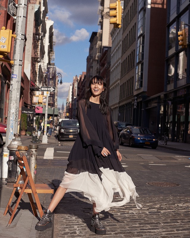 A young Asian woman stands in a black and white dress on a city street, with buildings and traffic lights in the background, medium shot.