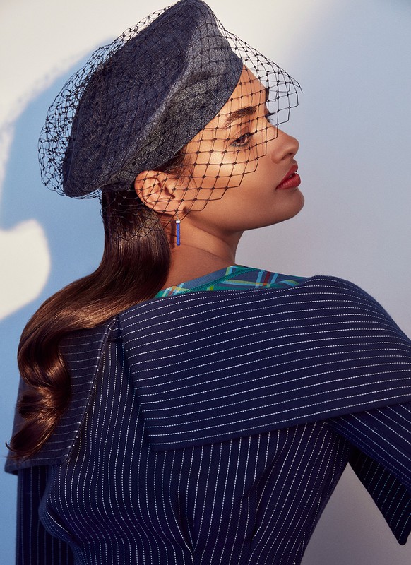 Profile shot of a woman in a pinstripe jacket and a black net fascinator against a light background. Studio lit fashion photograph.