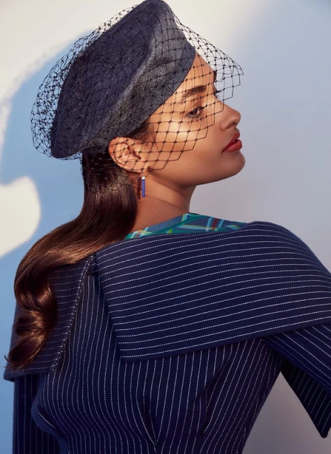 Profile shot of a woman in a pinstripe jacket and a black net fascinator against a light background. Studio lit fashion photograph.