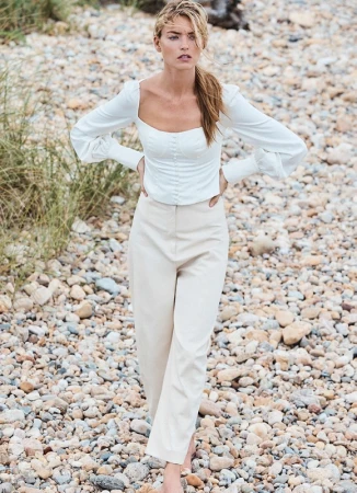 Model with a ponytail wearing a white long-sleeve top and light trousers on a rocky beach