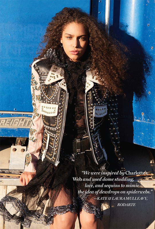Young woman with curly hair in a studded leather jacket and lace dress, standing in front of a blue truck trailer.