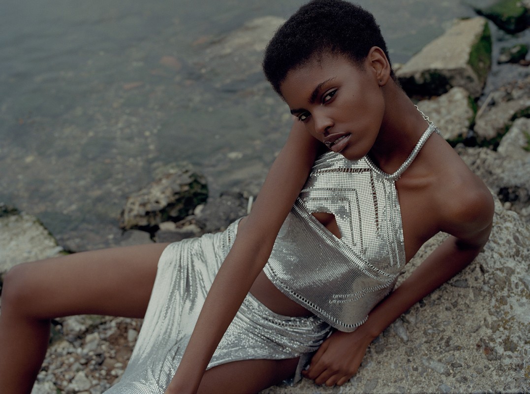 Adult female model with short hair in metallic silver crop top and skirt lying on rocky shoreline near grey water, looking at camera.