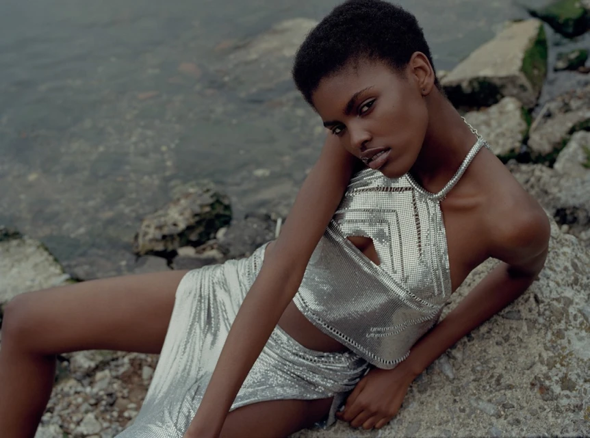 Adult female model with short hair in metallic silver crop top and skirt lying on rocky shoreline near grey water, looking at camera.