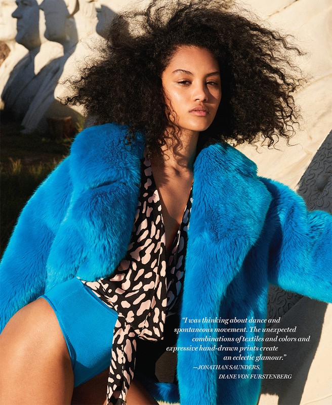 Fashion shot of a young woman with curly hair wearing a bright blue coat and patterned top, posing outdoors in front of stone structures.