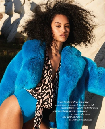Fashion shot of a young woman with curly hair wearing a bright blue coat and patterned top, posing outdoors in front of stone structures.