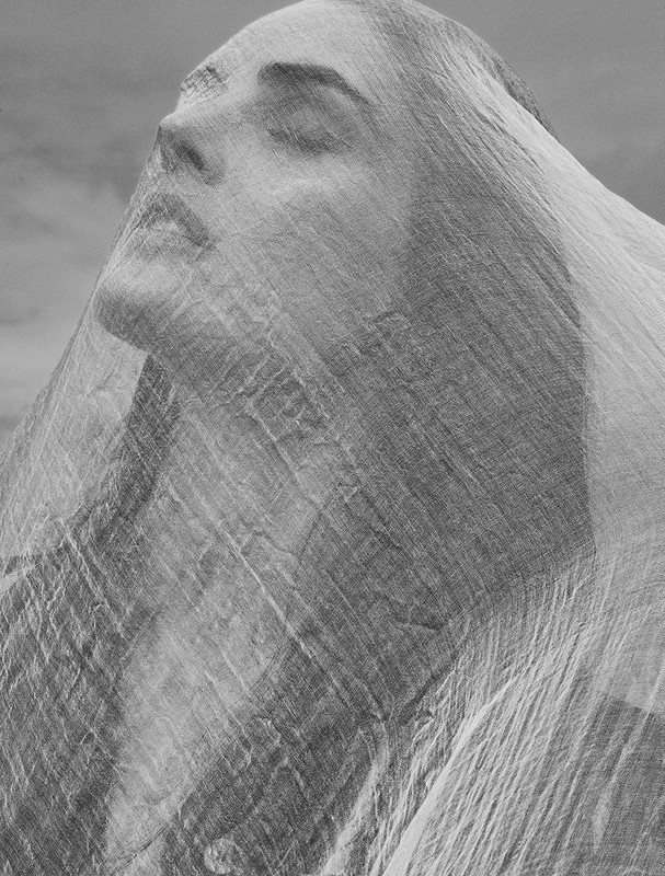 Monochrome portrait of an adult female with her eyes closed, draped in white fabric against a cloudy sky backdrop. Soft lighting.