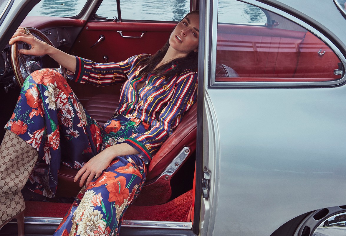 Woman in floral pants sits in a vintage silver car with a red interior, posing with her hand on the steering wheel in daylight.