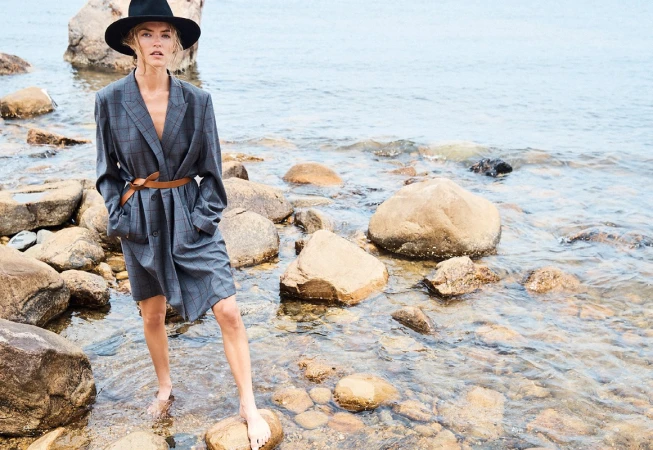 Model standing on wet rocks wearing a grey plaid blazer dress, brown leather belt, and black wide-brim hat by Campbell Ritchie.