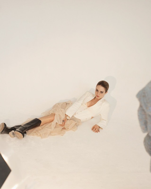 Adult woman in off-white jacket, beige tulle skirt and black boots reclines on a white background in a studio shot.