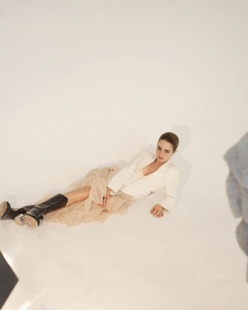 Adult woman in off-white jacket, beige tulle skirt and black boots reclines on a white background in a studio shot.