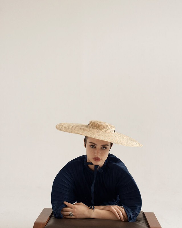 Portrait of a woman in a navy top and a wide-brimmed straw hat, seated with her arms crossed, set against a soft white background.