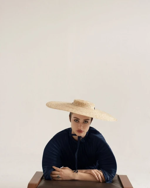 Portrait of a woman in a navy top and a wide-brimmed straw hat, seated with her arms crossed, set against a soft white background.