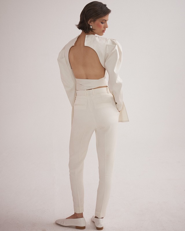 Back view of a woman posing in a cream-colored, open-back, puff-sleeve top and matching pants, with white flats against a plain background.