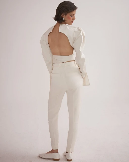 Back view of a woman posing in a cream-colored, open-back, puff-sleeve top and matching pants, with white flats against a plain background.