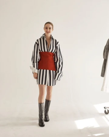 Model in vertical striped shirt dress with a red waist wrap and black boots in a studio setting