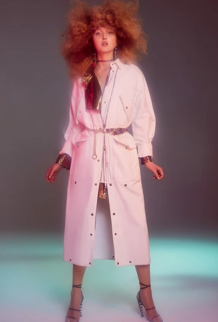 Full-length shot of a woman with voluminous red hair, wearing a white jacket dress, strappy heels, and statement jewelry on a gradient background.