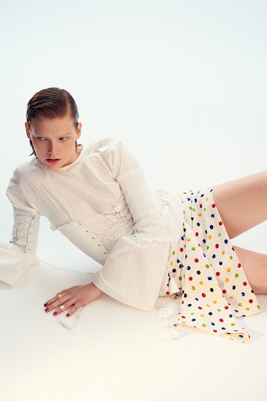 A fair-skinned woman in a white dress with colorful polka dots poses in a white studio, showcasing fashion and editorial style.