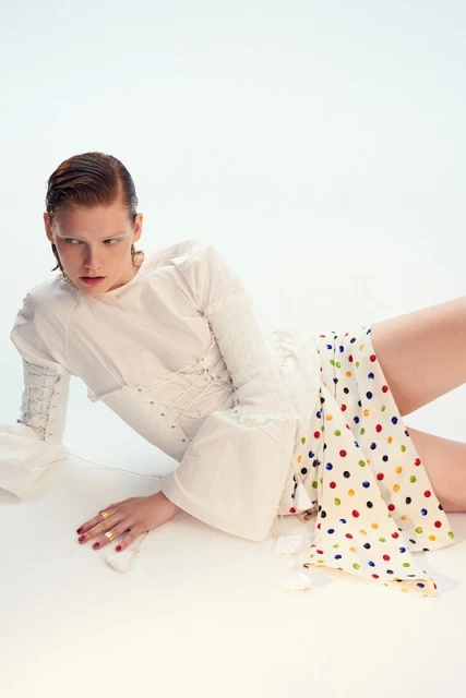 A fair-skinned woman in a white dress with colorful polka dots poses in a white studio, showcasing fashion and editorial style.